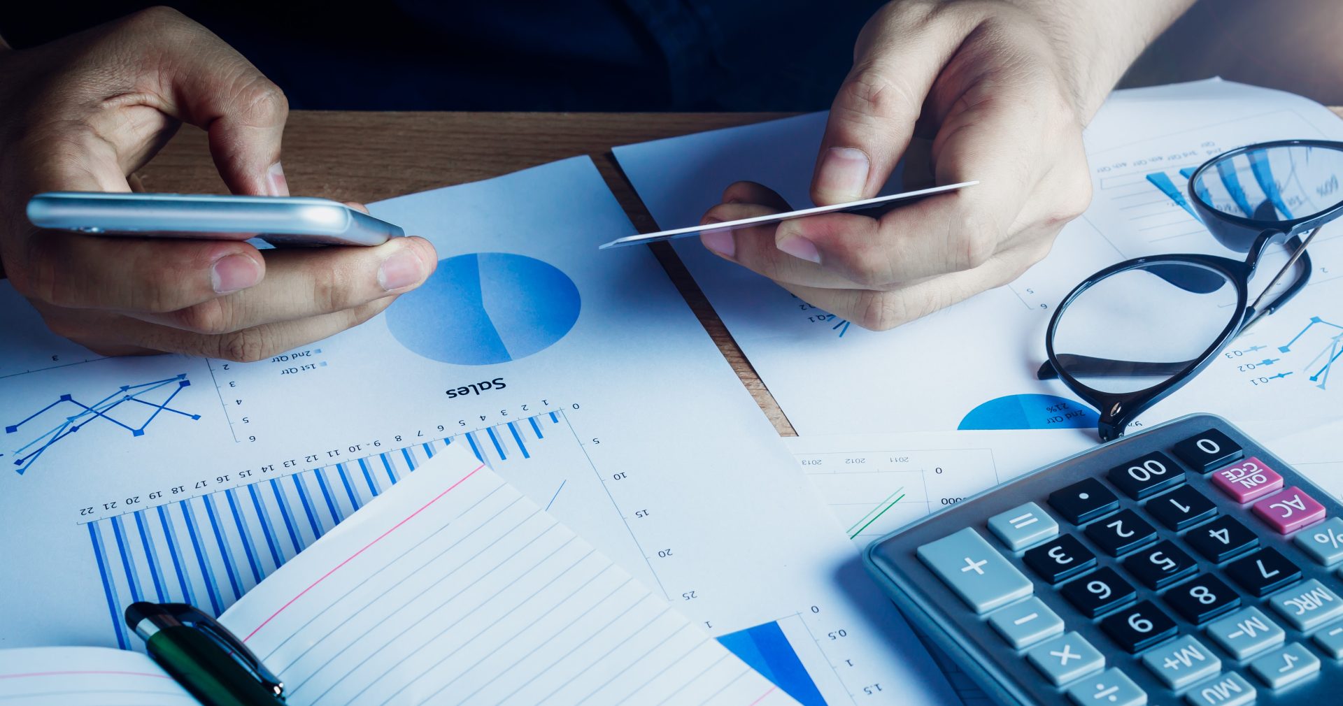 PIX para Empresas: quais as Vantagens para os Pequenos Negócios? Homem com cartão e celular, em uma mesa cheia de planilhas, realizando um pix para empresas